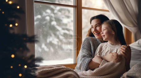 Téléchargez les photos : Joyeux famille aimante. Mère et fille s'embrassent et profitent de la nature hivernale dans la fenêtre. - en image libre de droit