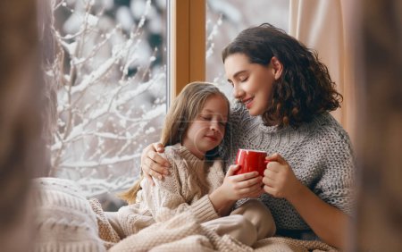 Téléchargez les photos : Joyeux famille aimante. Mère et fille s'embrassent et boivent des boissons chaudes en profitant de la nature hivernale dans la fenêtre. - en image libre de droit