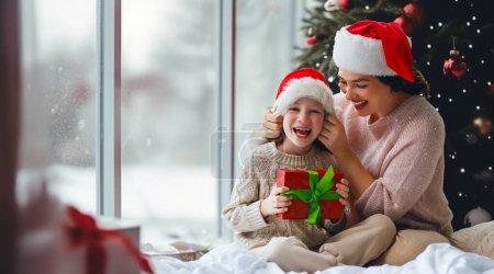 Photo for Merry Christmas and Happy Holidays. Cheerful mom and her cute daughter girl exchanging gifts. Parent and little child having fun near tree indoors. Morning Xmas. - Royalty Free Image