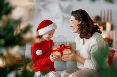 Photo for Merry Christmas and Happy Holidays. Cheerful mom and her cute daughter girl exchanging gifts. Parent and little child having fun near tree indoors. Loving family with presents in room. - Royalty Free Image