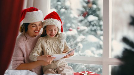 Téléchargez les photos : Joyeux Noël et Joyeuses Fêtes. Joyeux maman et sa fille mignonne fille lecture d'un livre. Parent et petit enfant s'amusent près de l'arbre à l'intérieur. Matin Noël. - en image libre de droit