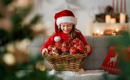 Foto de Feliz Navidad y Felices Fiestas. Linda niña está decorando el árbol en el interior. La mañana antes de Navidad. Retrato niño de cerca. - Imagen libre de derechos