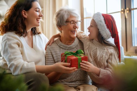 Photo for Merry Christmas and Happy Holidays. Cheerful kid presenting gifts to mom and granny. Parents and little child having fun near tree indoors. Loving family with presents in room. - Royalty Free Image