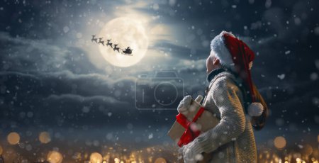 Photo for Merry Christmas. Cute little child with xmas present. Santa Claus flying in his sleigh against moon sky. Happy kid enjoy the holiday. Portrait of girl with gifts on dark background. - Royalty Free Image