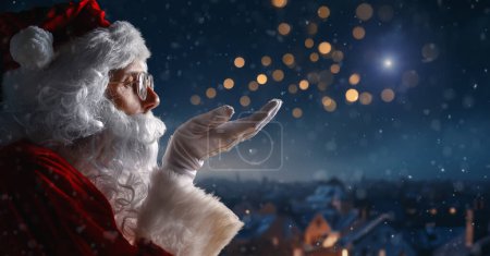 Foto de Feliz Navidad y felices fiestas. Santa Claus soplando nieve y mirando a la ciudad nocturna. - Imagen libre de derechos