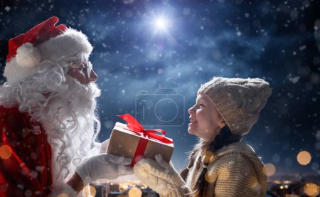 Téléchargez les photos : Joyeux Noël et bonnes vacances. Petite fille mignonne et Père Noël avec un cadeau. Concept légende de Noël. - en image libre de droit