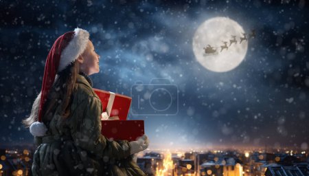 Photo for Merry Christmas. Cute little child with xmas present. Santa Claus flying in his sleigh against moon sky. Happy kid enjoy the holiday. Portrait of girl with gifts on dark background. - Royalty Free Image