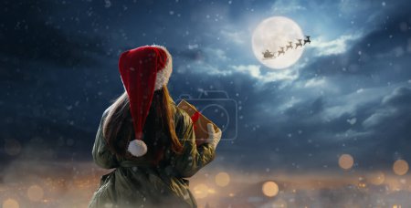 Téléchargez les photos : Joyeux Noël. Petit enfant mignon avec cadeau de Noël. Le Père Noël volant dans son traîneau contre le ciel lunaire. Heureux gamin profiter des vacances. Portrait de fille avec des cadeaux sur fond sombre. - en image libre de droit