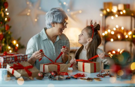 Photo for Happy Holidays. Cheerful grandmother and her cute granddaughter girl preparing for Christmas. People wrapping gifts, decorating home. Loving family with presents in room. - Royalty Free Image