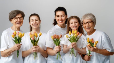 Foto de ¡Feliz día de las mujeres! Las hijas de los niños están felicitando a mamá y abuelas dándoles tulipanes de flores. Grannys, mamá y niñas sonriendo sobre fondo gris claro. Vacaciones familiares y unión. - Imagen libre de derechos
