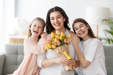 Téléchargez les photos : Bonne fête des mères. Les filles des enfants félicitent maman et lui donnent des fleurs. Maman et les filles souriant et étreignant. Vacances en famille et convivialité. - en image libre de droit
