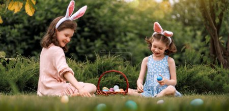 Photo for Beautiful children with painting eggs outdoors. Happy family celebrating Easter in nature. Cute little girls are wearing bunny ears. - Royalty Free Image