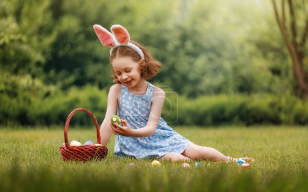 Photo for Beautiful child with painting eggs outdoors. Happy family celebrating Easter in nature. Cute little girl is wearing bunny ears. - Royalty Free Image