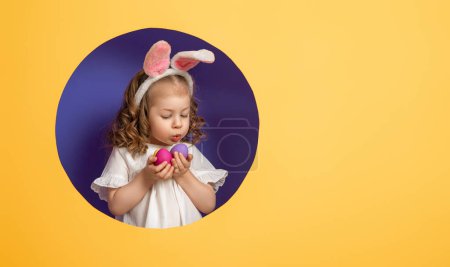 Téléchargez les photos : Mignon petit enfant portant des oreilles de lapin le jour de Pâques. Fille avec des œufs peints sur fond violet vif et jaune
. - en image libre de droit