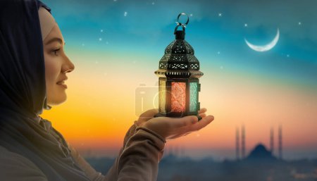 Foto de Mujer sostiene linterna árabe ornamental con vela encendida que brilla en el fondo de la mezquita de la noche. Tarjeta de felicitación festiva, invitación para el mes sagrado musulmán Ramadán Kareem. - Imagen libre de derechos