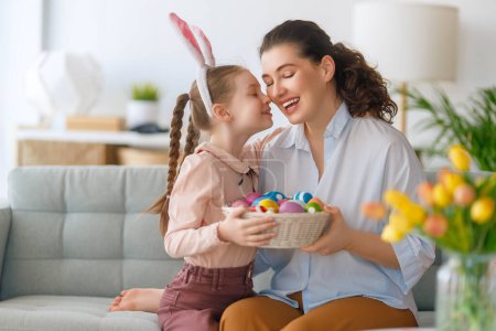 Foto de ¡Felices vacaciones! Madre y su hija pintando huevos. Familia celebrando la Pascua. Linda niña está usando orejas de conejo. - Imagen libre de derechos