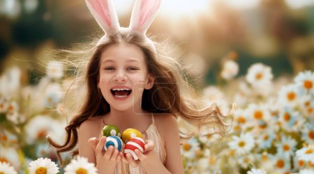 Téléchargez les photos : Belle enfant avec la peinture des œufs à l'extérieur. Joyeuse famille célébrant Pâques dans la nature. Petite fille mignonne porte des oreilles de lapin. - en image libre de droit