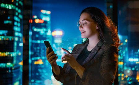 Téléchargez les photos : Jeune femme d'affaires utilisant le téléphone près de la fenêtre le soir. - en image libre de droit