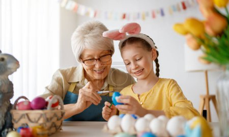 Téléchargez les photos : Une grand-mère et sa petite-fille peignant des œufs. Bonne famille se préparant pour Pâques. Petite fille mignonne portant des oreilles de lapin. - en image libre de droit