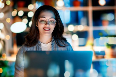 Téléchargez les photos : Jeune femme d'affaires travaillant sur ordinateur portable près de la fenêtre dans la soirée. - en image libre de droit