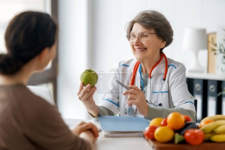 Téléchargez les photos : Heureux médecin travaille dans le bureau de médecine. Une femme parle à un patient de l'importance de manger des fruits et légumes. - en image libre de droit