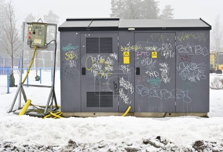 An electrical power supply box is covered with various graffiti tags, standing in a snow-covered landscape with fog in the background