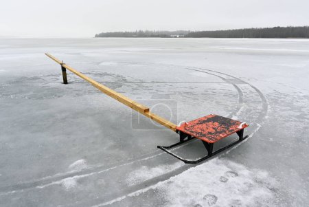 Ein trostloser zugefrorener See mit einem einsamen roten Schlitten, der an einer langen Holzplanke befestigt ist und einen starken Kontrast bildet