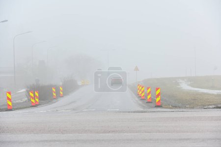 Téléchargez les photos : Une voiture solitaire roule sur une route désolée et brumeuse flanquée de panneaux de signalisation routière, avec un environnement à peine visible en raison du brouillard épais - en image libre de droit