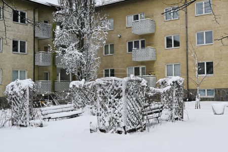 A residential area with trees and a bench covered in snow in front of a yellow apartment building with balconies