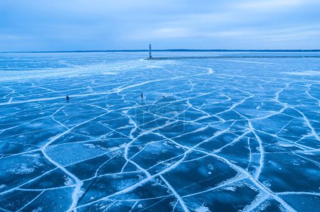 Téléchargez les photos : Lac gelé, glace bleue avec fissures. Pêcheurs pêchent, un phare avec un barrage en arrière-plan - en image libre de droit