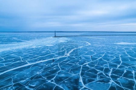 Téléchargez les photos : Lac gelé, glace bleue avec fissures. Pêcheurs pêchent, un phare avec un barrage en arrière-plan - en image libre de droit