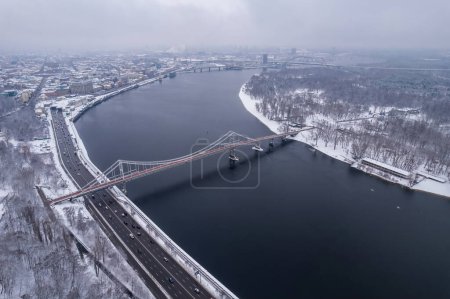 Photo for Aerial drone view. Pedestrian bridge over the frozen Dnieper River in Kiev. Cloudy frosty winter morning, frosty pattern on ice. - Royalty Free Image