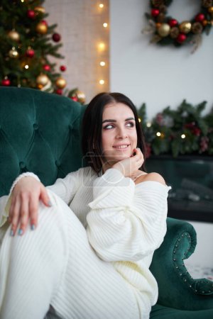 Foto de Feliz joven soñadora sentada cerca del árbol de Navidad en jersey blanco. Sonriente joven mujer disfrutando de vacaciones de invierno de año nuevo en casa, soñando o imaginando un futuro alegre. - Imagen libre de derechos