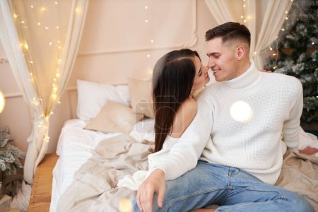 Photo for Passionate romantic couple spending time before New Year at home with garlands. Sexy young woman and handsome man lying on bed in sweaters and hugging. Tender couple together - Royalty Free Image
