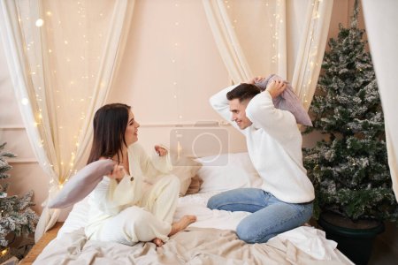 Téléchargez les photos : Joyeux jeune couple charmant s'amuser, se battre avec des oreillers sur le lit dans la chambre à coucher festivement décorée du Nouvel An, couple profitant de la compagnie de l'autre, passer du temps à la maison pendant Noël - en image libre de droit