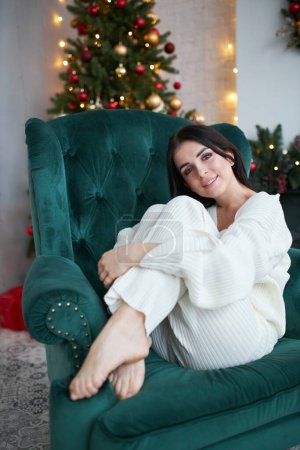 Photo for Happy dreamy young woman sitting near christmas tree in white sweater. Smiling young female enjoying new year winter holiday at home, dreaming or imagining a joyful future. - Royalty Free Image