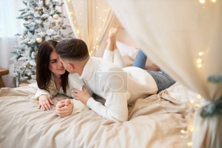 Foto de Sonriendo encantadora novia sintiendo felicidad estar con su novio y comunicarse entre sí, pareja romántica positiva relajarse en la cama durante las vacaciones de invierno en el acogedor interior del hogar - Imagen libre de derechos