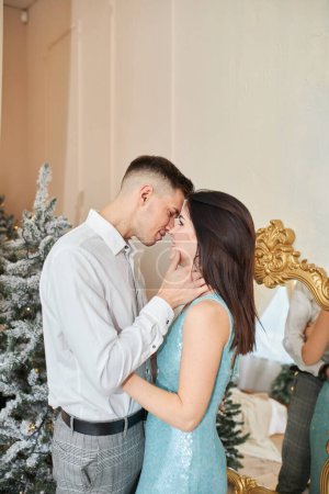 Foto de Joven dulce bonita pareja enamorada besándose en víspera de Navidad cerca del árbol con regalos en casa en diciembre
. - Imagen libre de derechos