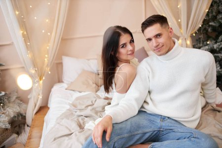 Photo for Passionate romantic couple spending time before New Year at home with garlands. Sexy young woman and handsome man lying on bed in sweaters and hugging. Tender couple together - Royalty Free Image