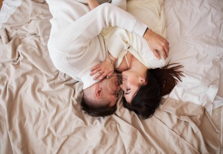 Photo for Top view of passionate romantic couple spending time before New Year at home with garlands. Sexy young woman and handsome man lying on bed in sweaters and hugging. Tender couple together - Royalty Free Image