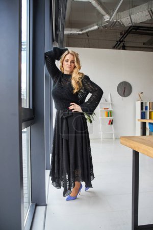 Téléchargez les photos : Young cute blonde posing in a studio near the window dressed in an evening black dress - en image libre de droit