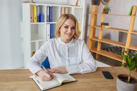 Photo for Portrait of a happy business woman writing down notes while sitting at table in the office. - Royalty Free Image