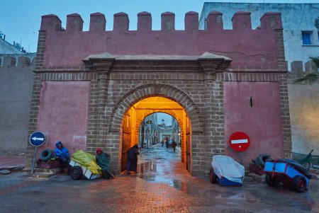 Foto de ESSAOUIRA, MOROCCO. 13 th February, 2017: Ancient walls of the medina with palm trees, merchants hurrying on business, after the rain - Imagen libre de derechos