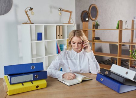Foto de A young girl with long blond hair sits at a desk in the office, works on a computer and writes in a diary. - Imagen libre de derechos