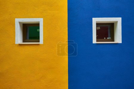 Foto de Hermosa fachada colorida de la casa en la isla de Burano, norte de Italia. Mitad naranja mitad azul pared de la casa con una puerta amarilla y dos ventanas con plantas en ella - Imagen libre de derechos