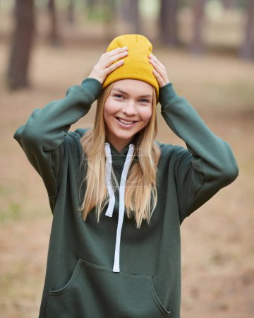 Foto de Atractiva mujer rubia de ojos azules caminar por el parque. Chica usar sudadera con capucha verde y sombrero amarillo, mirada feliz y sonrisas. Retrato de una joven alegre disfrutando en el parque de otoño. - Imagen libre de derechos