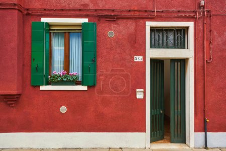 Téléchargez les photos : Porte et fenêtre avec des fleurs sur la façade rouge de la maison. Architecture colorée sur l'île de Burano, Venise, Italie. - en image libre de droit