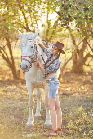 Foto de Feliz hermosa joven vaquera en sombrero de pie y abrazando a su caballo - Imagen libre de derechos