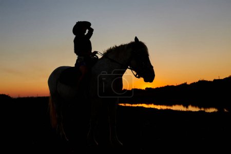 Foto de Mujer a caballo montando a caballo galopando con el sol naciente rojo en el horizonte. Hermoso fondo de cabecera colorido atardecer con silueta de equino y niñas. - Imagen libre de derechos