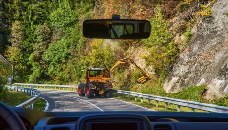 Foto de Un tractor con una recortadora grande en una grúa mecánica recorta los arbustos en una carretera de montaña en los Alpes. - Imagen libre de derechos